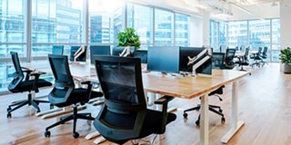 A modern office space with tall widows, bright lights, and office chairs surrounding work desks with built-in monitors