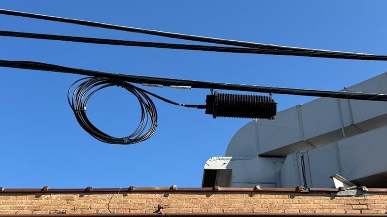A black fiber closure with coiled fiber cables hang from a utility wire with leaves in the background