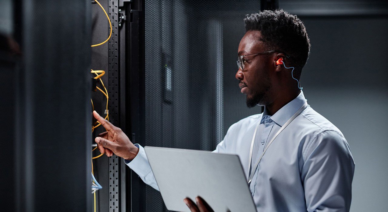 A man dressed in a shirt and tie is positioned in front of a server while holding a laptop.
