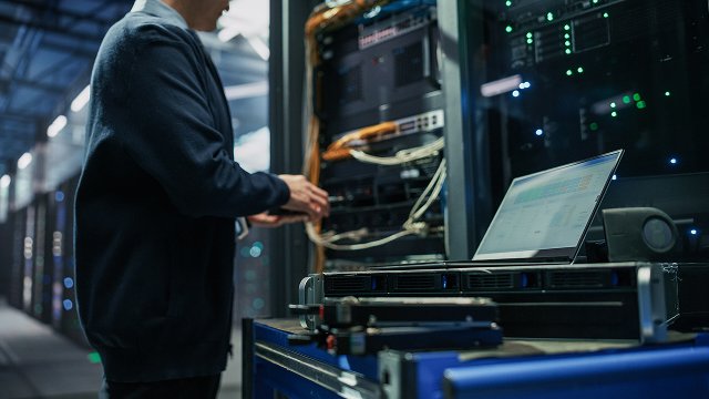 Low Angle View of Male IT Specialist Mounting Hardware Unit Into Server Rack While Using Laptop for Diagnostics 