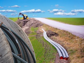 Trenching equipment digs a trench to lay fiber optic cable, which is shown on a wooden reel, with three pieces of conduit alongside a rural road