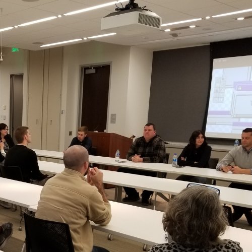 Table with panelists in front of room and tables with male and female adults listening