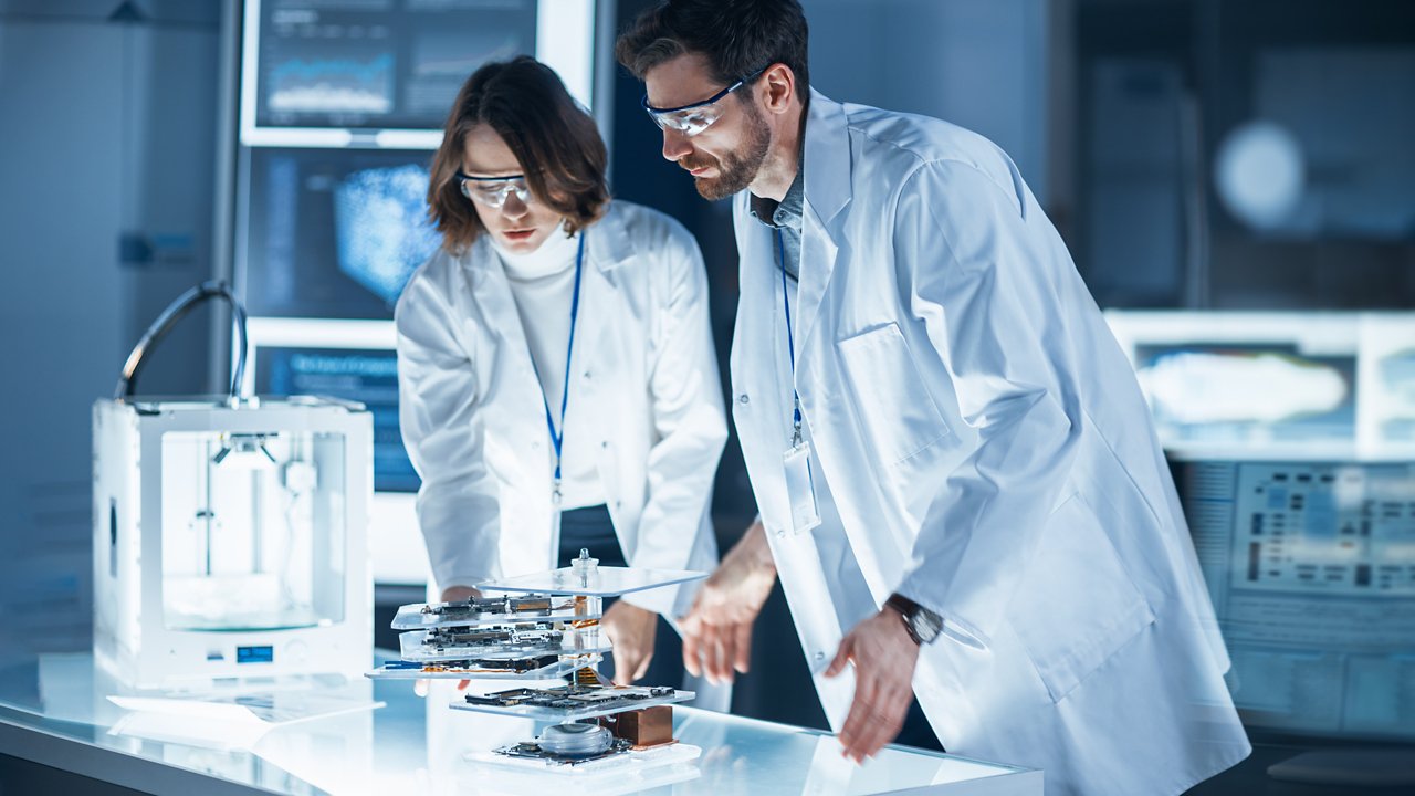 Diverse International Team of Industrial Scientists and Engineers Wearing White Coats Working on Heavy Machinery Design in Research Laboratory. Professionals Using 3D Printer, Computers and Microscope