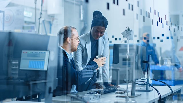 A man and women sitting in a lab with a glass front looking at a product in man’s hand
