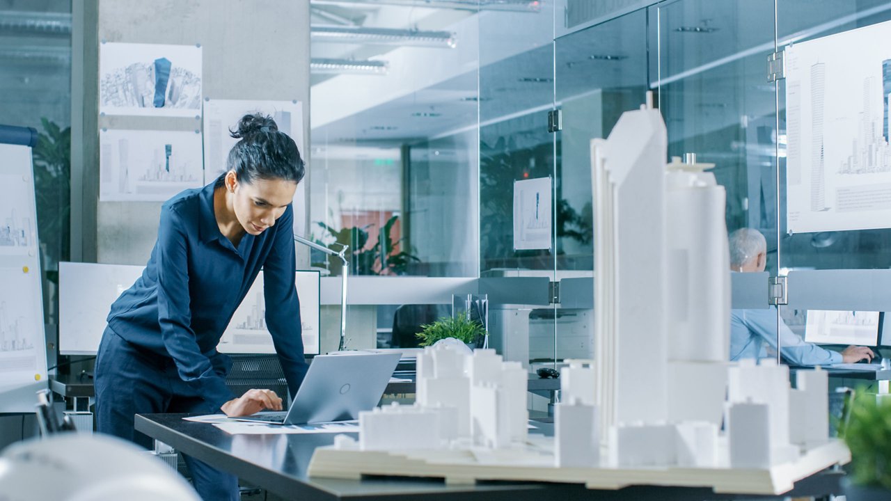Female Architectural Designer Works on a Laptop,  Engineering New Building Model for the Urban Planning Project. Clean Minimalistic Office, Concrete Walls Covered by Blueprints and Documents.