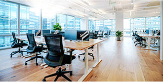 Open office area with large windows and rows of tables with chairs and monitors