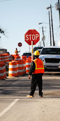 a worker in safety gear holds a stop sign to direct traffic through a road construction zone, with vehicles waiting to pass