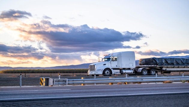 Industrial long hauler big rig white semi truck with chrome parts transporting fastened by slings covered commercial cargo on flat bed semi trailer running on the straight highway road