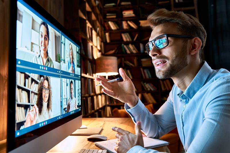 A bearded man holding a cell phone sitting at a computer on a video call