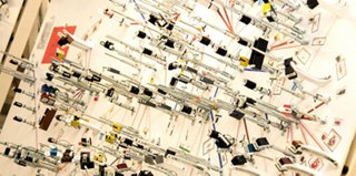 Wall-mounted display of multiple metal linkage assemblies, cables, and industrial components arranged across a whiteboard with labeled diagrams, illustrating a variety of mechanical or wiring configurations.