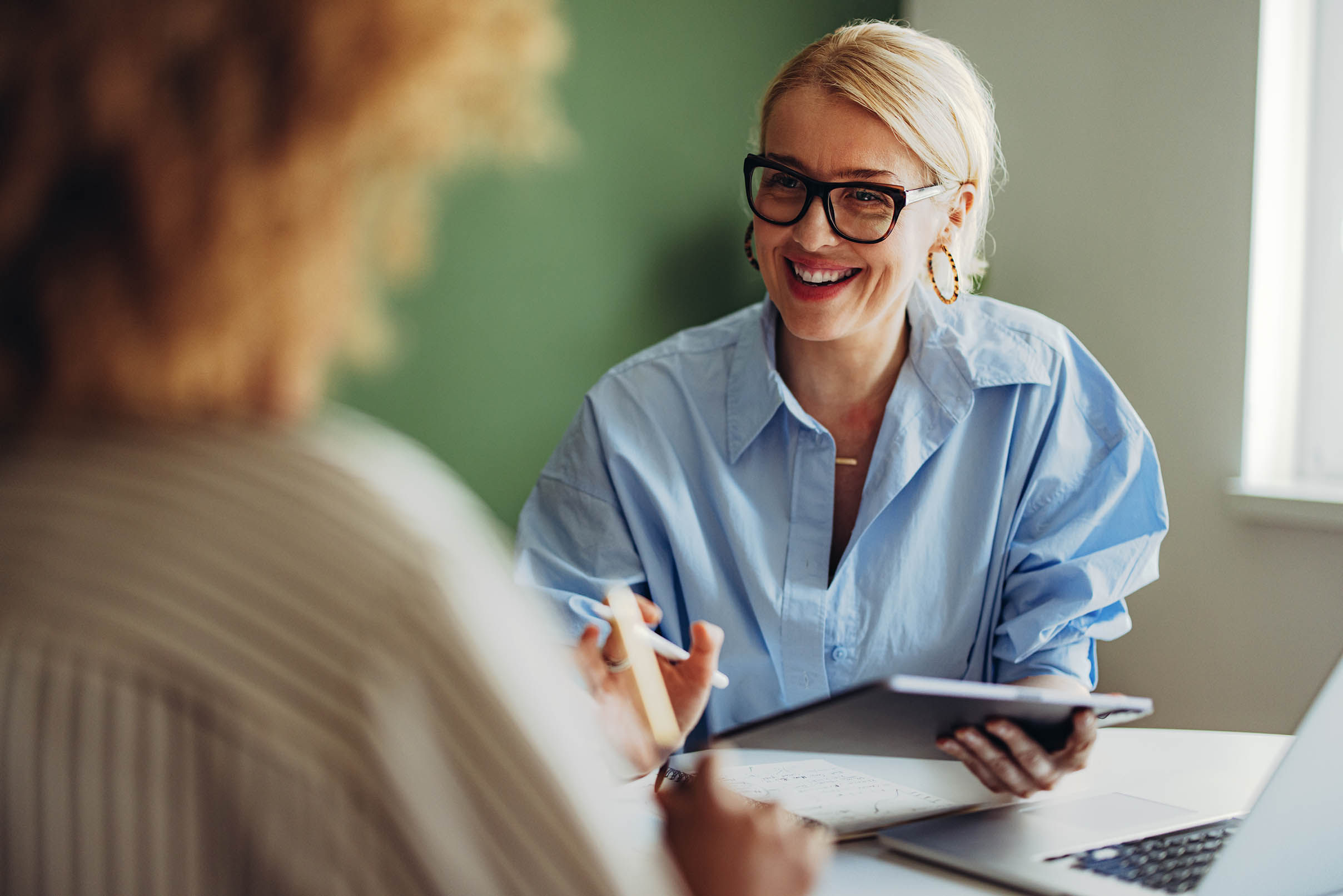 Two women conversing in corporate setting with laptop and tablet