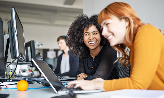 Two young women working together on laptop with male colleague in background. Two female colleagues in office working together.
