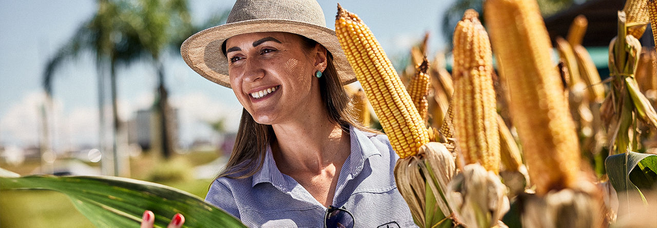 Mulher com camisa e chapéu da Pioneer sorrindo e segurando uma espiga de milho na lavoura