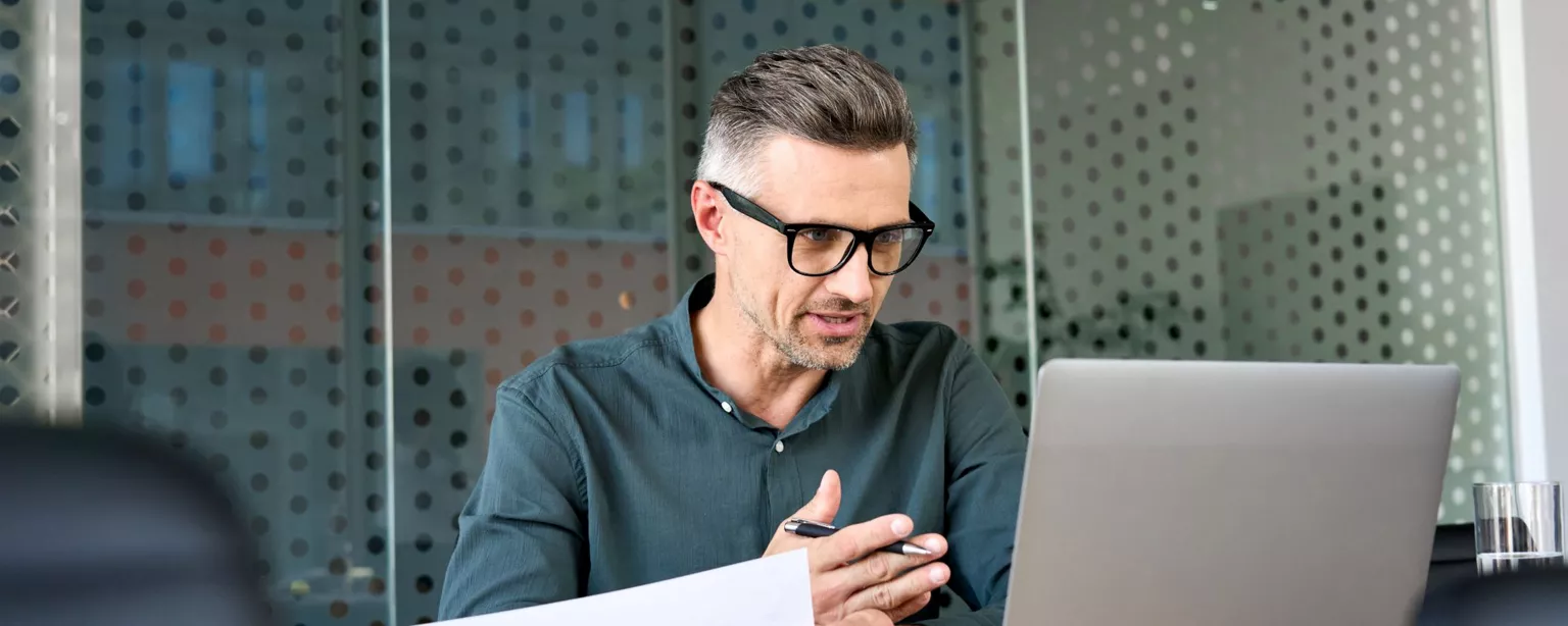 A man with glasses sits in a conference room and uses his laptop to conduct a virtual interview with a job candidate.