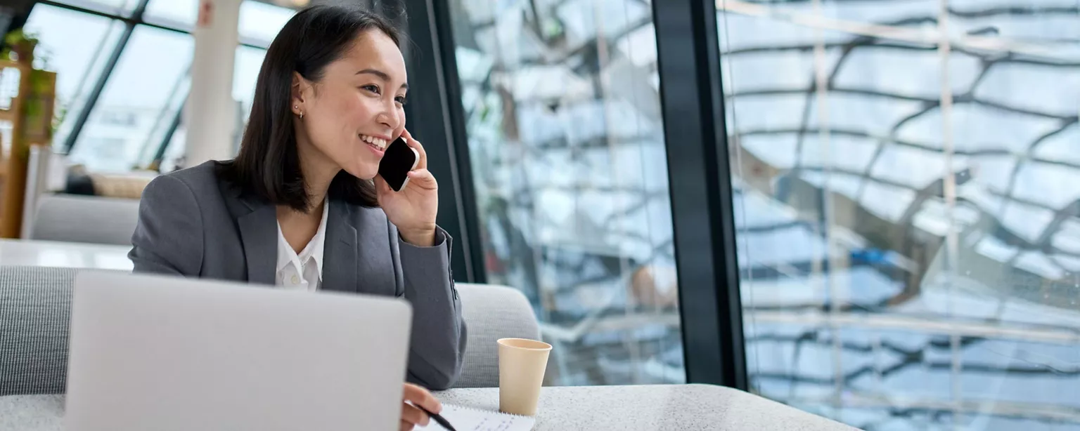 A professional woman in a business suit is smiling and taking notes while speaking on the phone, seated at a desk with a laptop and a coffee cup in an office setting.