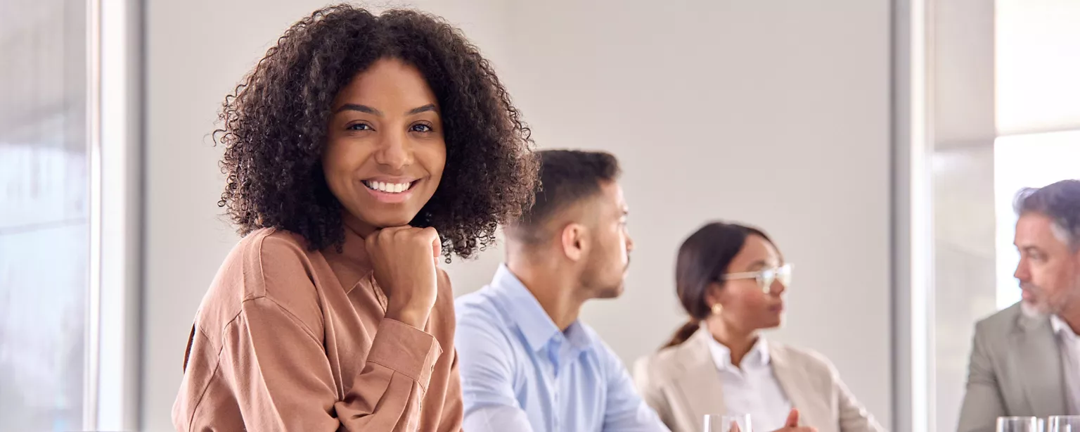 A smiling woman sits confidently at a meeting table <a href=