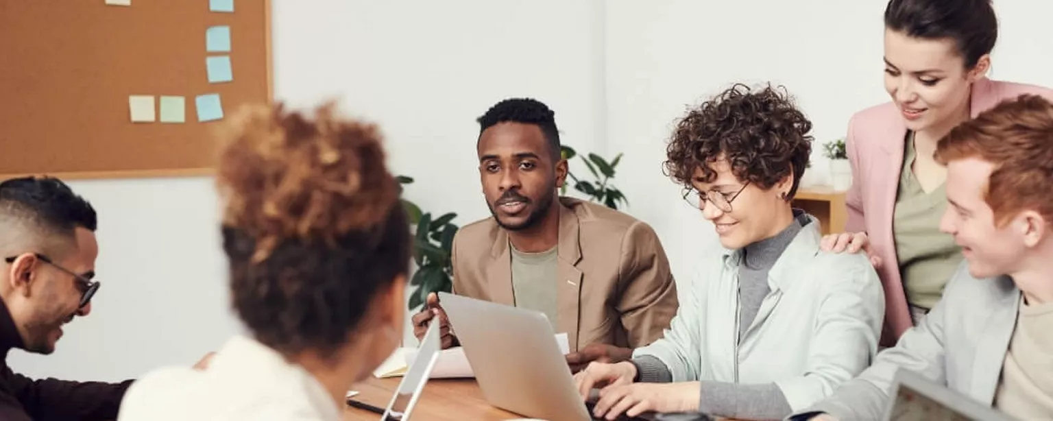 A diverse group of employees appear in a brainstorm, with a combination of laptops and notes on a tackboard