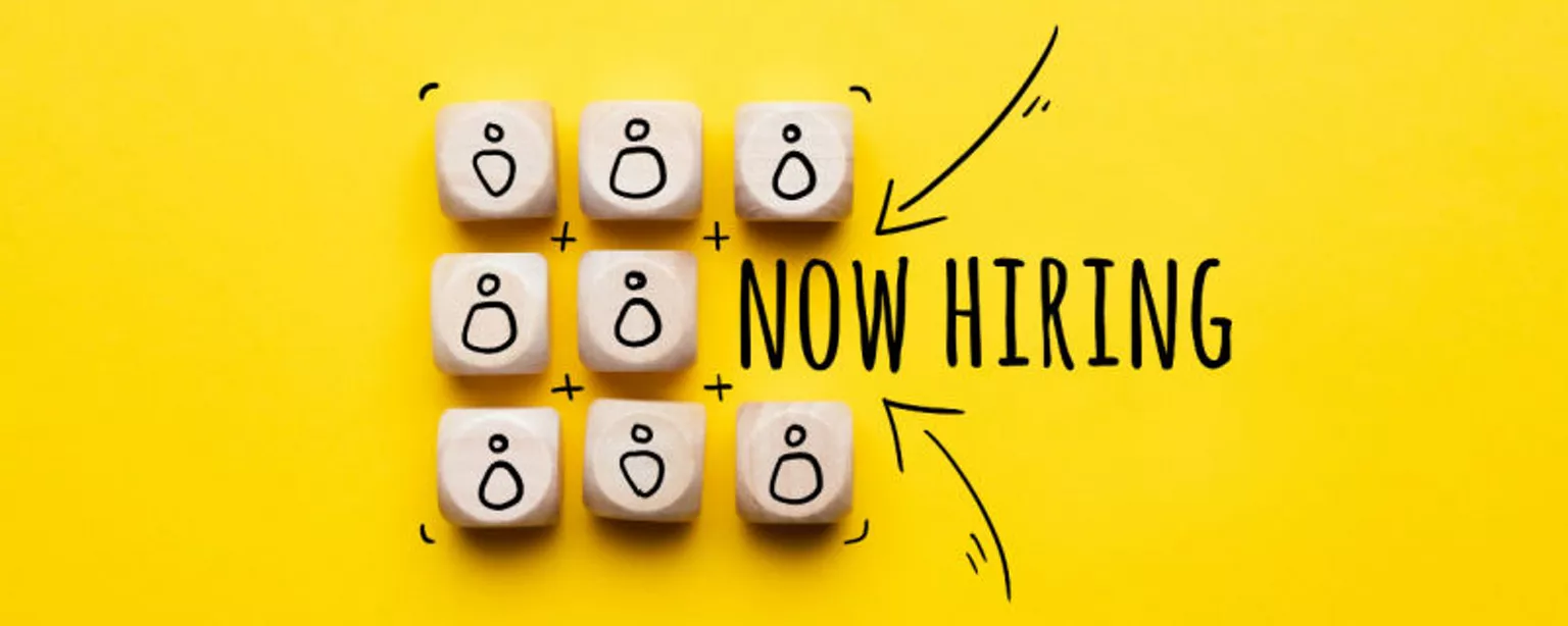 Small wooden blocks representing the workforce are displayed next to the words "Now Hiring" against a yellow background.