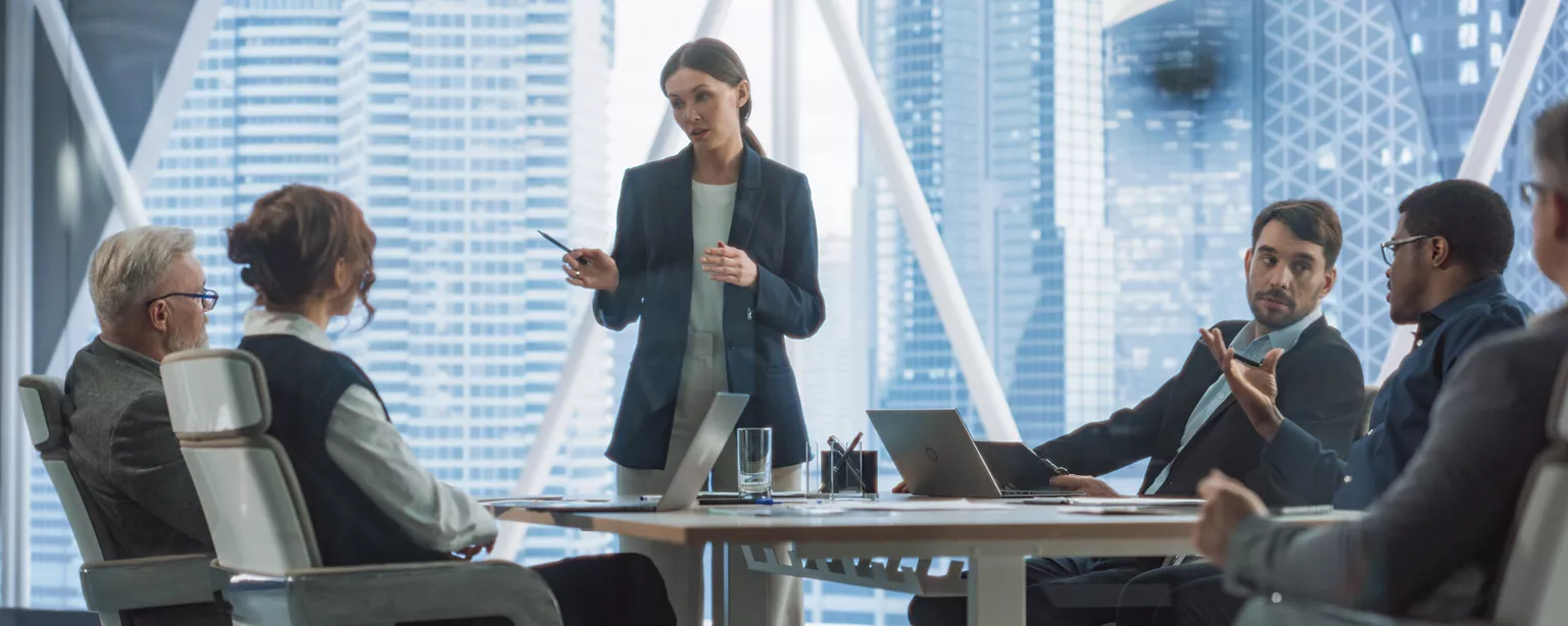 A female leads a Board Of Directors Meeting in a corporate office. The board is a diverse group of men and women around a conference table.