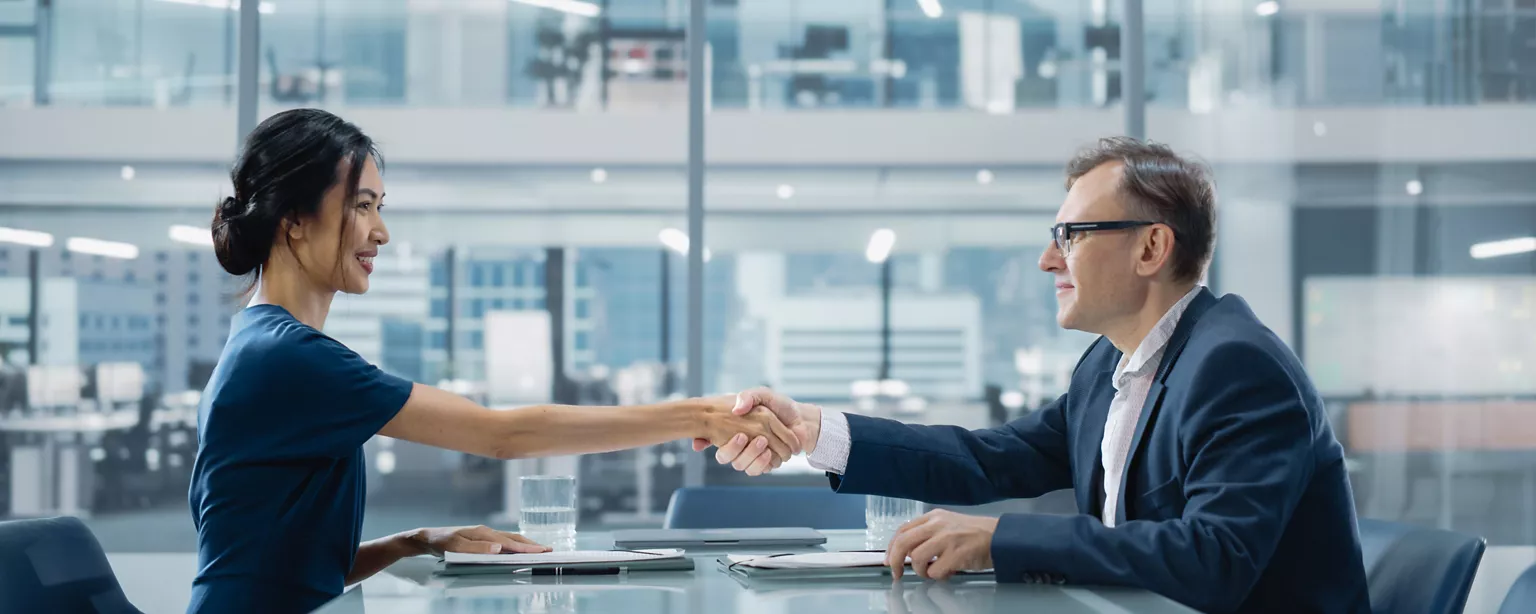 Two sharply dressed and smiling business executives, a woman and a man, shake hands across a table in a conference room in a modern office setting.