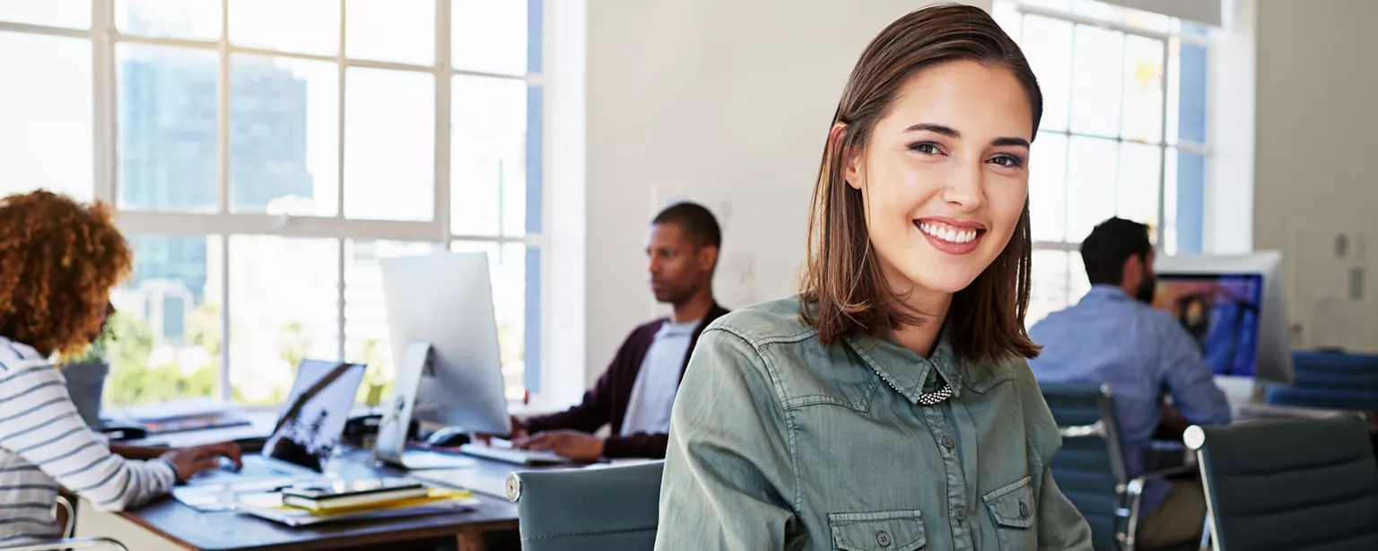 A smiling young professional is seated in an open office with colleagues working in the background.