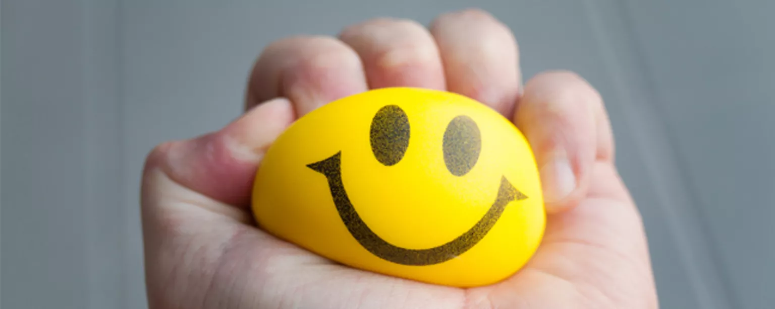 A hand squeezes a yellow ball with a smiley face imprinted on it.