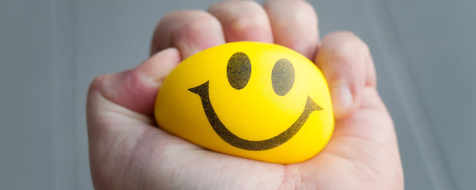 A hand squeezes a yellow ball with a smiley face imprinted on it.