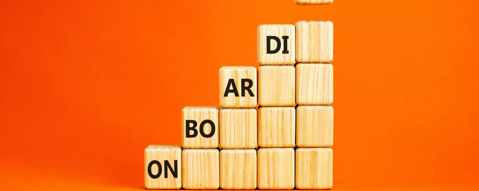 Wooden blocks set up like steps against an orange background with the word "Onboarding" staggered across the levels; a hand is placing a block with the letters "NG" on the top "step" of blocks.