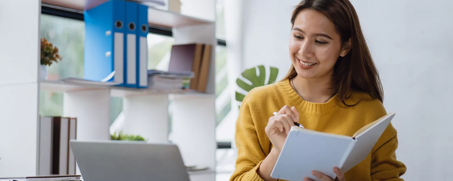 A woman in a yellow sweater smiles while taking notes from a laptop during a work or study session.