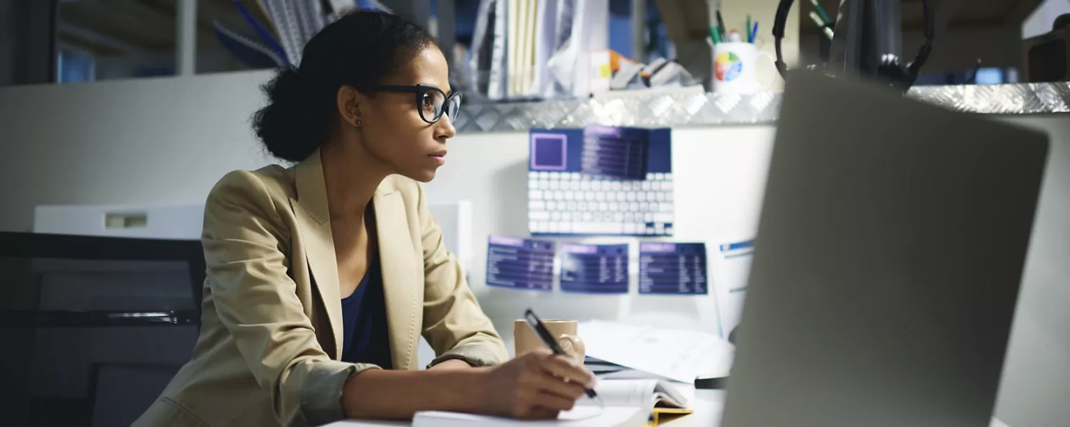 A focused professional wearing glasses works at her desk, taking notes while looking at a computer screen.