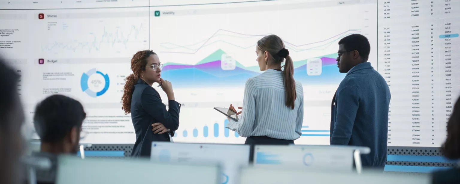 Three business professionals review financial data and analytics on a large digital screen during a strategic planning meeting.
