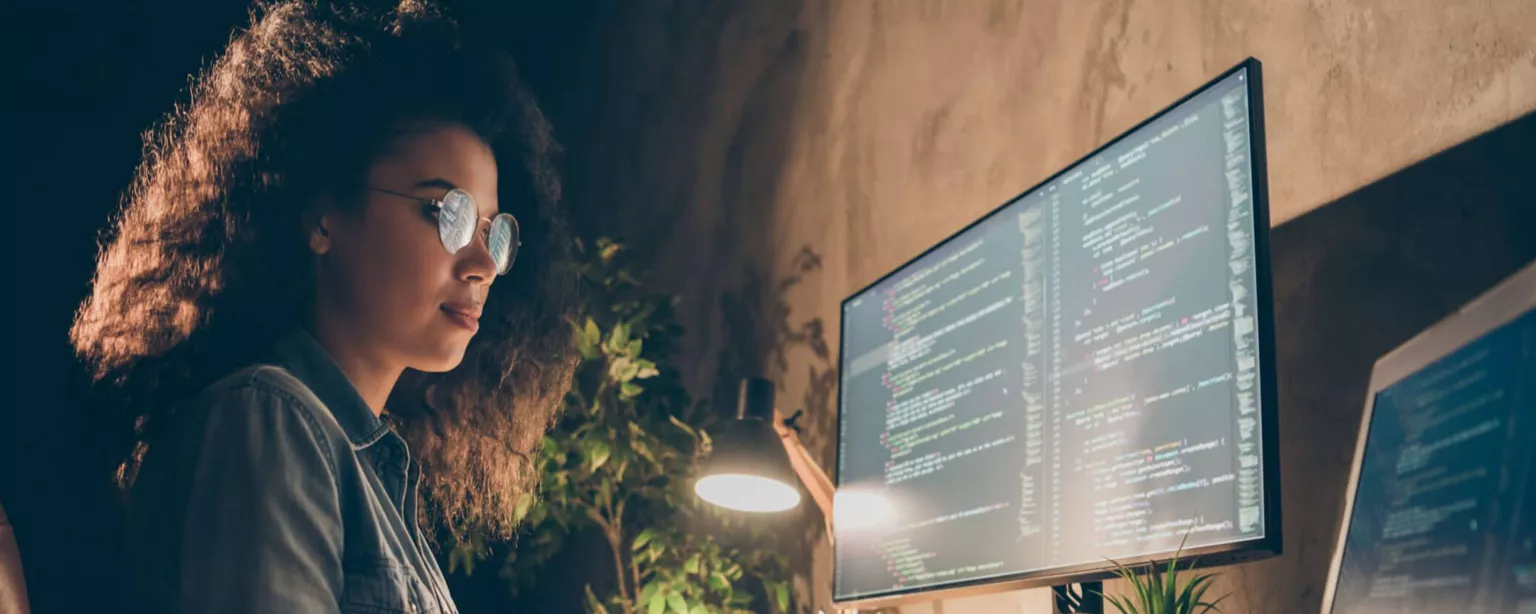 A young woman with glasses looks at computer code on her desktop and laptop screens.