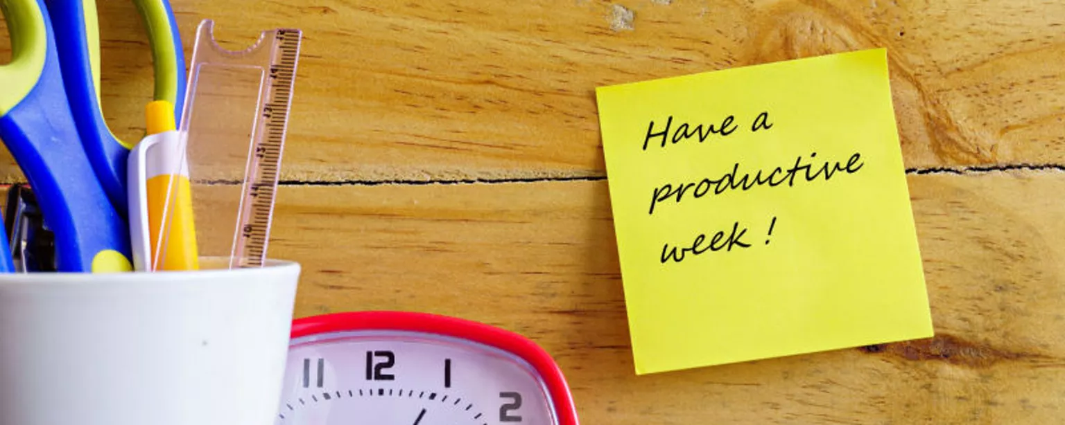A yellow sticky note affixed to a table near a clock and a cup with scissors and pens advises "Have a productive week!" 