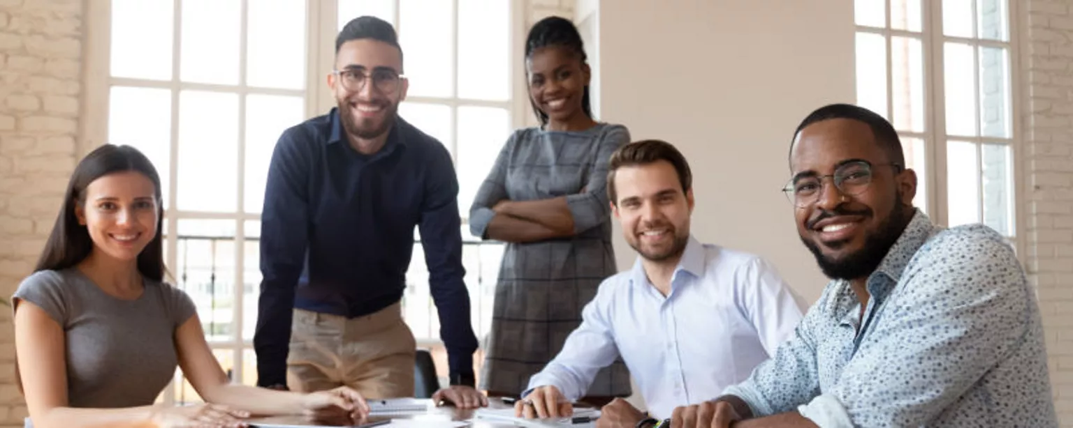 Five business colleagues gathered around a table in a modern office and smiling toward the camera.