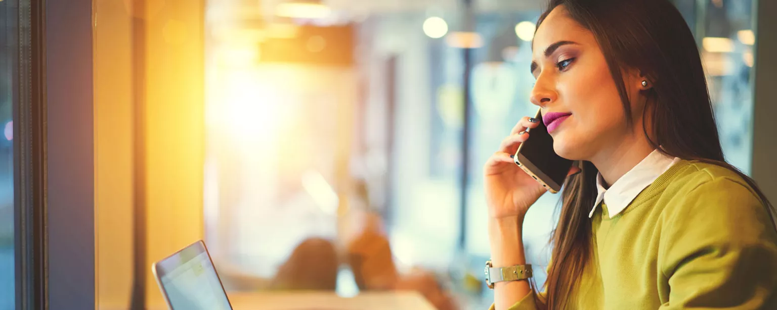 dark heaired woman in profile wearing green sweater taps on laptop while holding smartphone to her ear