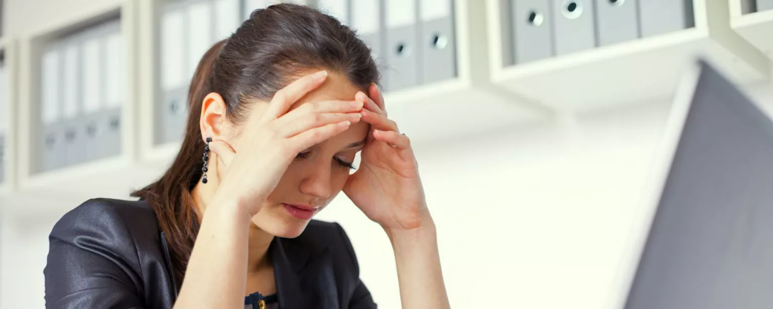 Young woman sitting a desk in front of a laptop, head in hands and eyes closed, looking stressed.