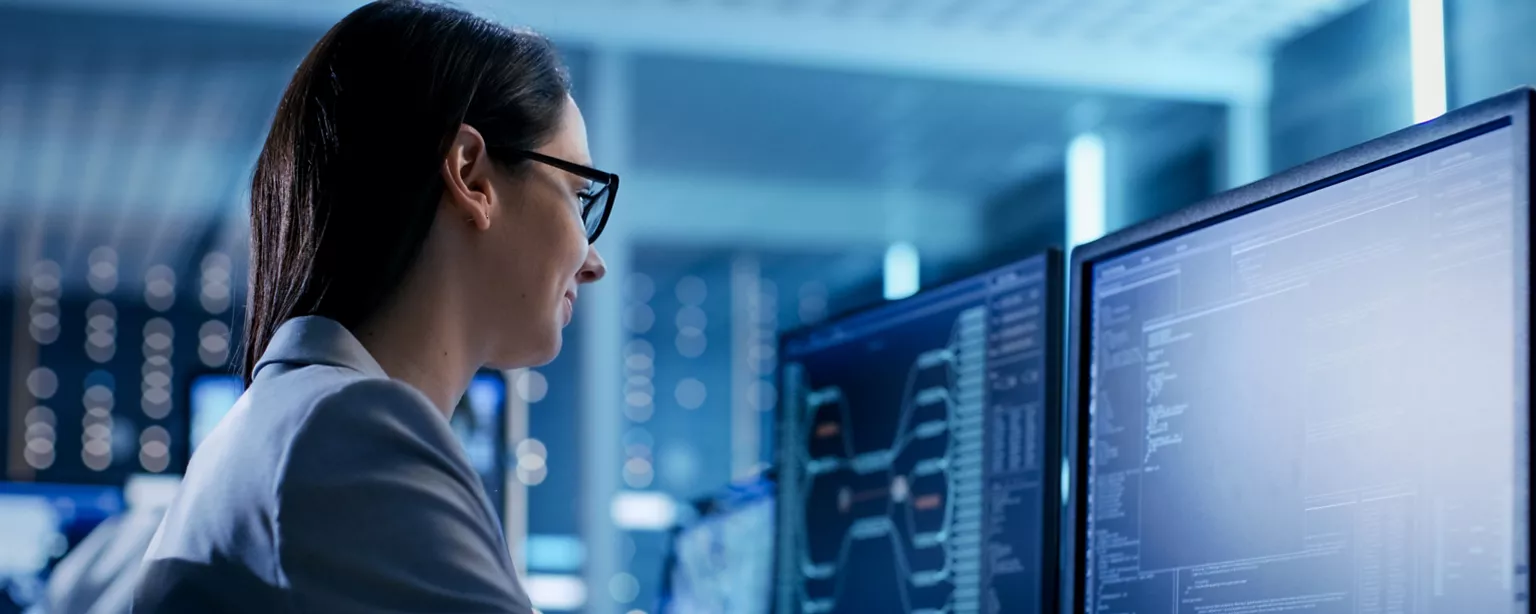 A woman, a cybersecurity professional, works in a security operations center. She is looking at data on two computer screens.