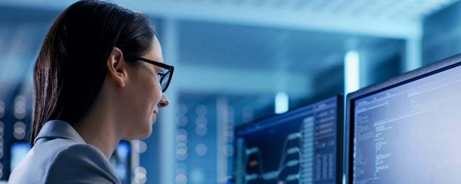 A woman, a cybersecurity professional, works in a security operations center. She is looking at data on two computer screens.