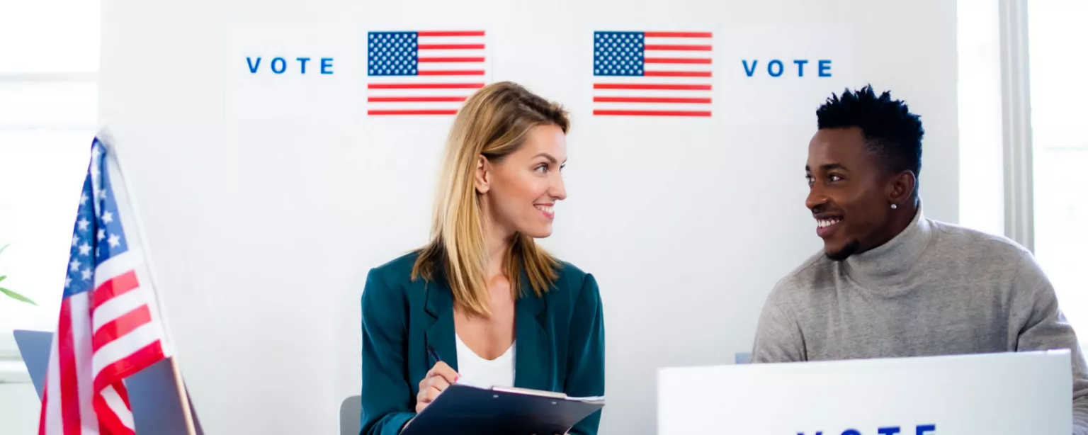 woman holding clipboard and man with laptop labeled "vote" seated behind a table with American flags and "vote on backdrop