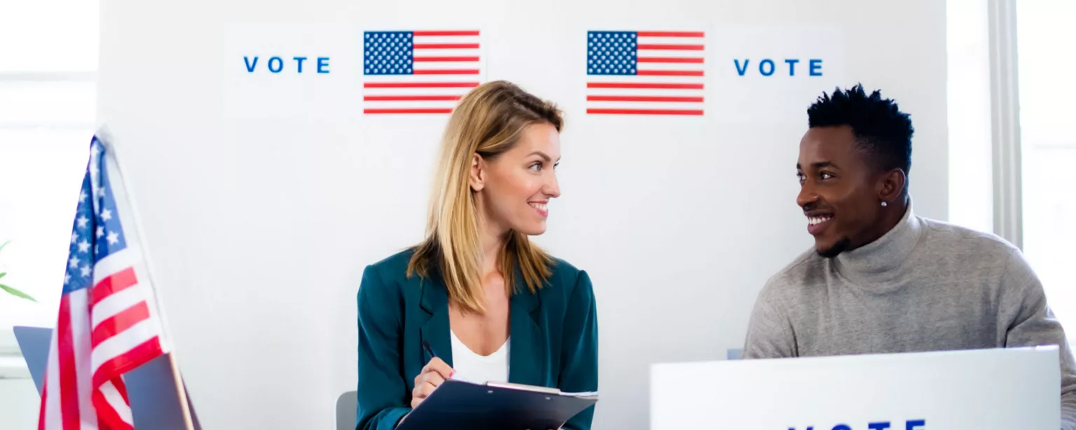 A woman holding a clipboard and a man with laptop labeled "vote" seated behind a table with American flags and "vote" on the backdrop.