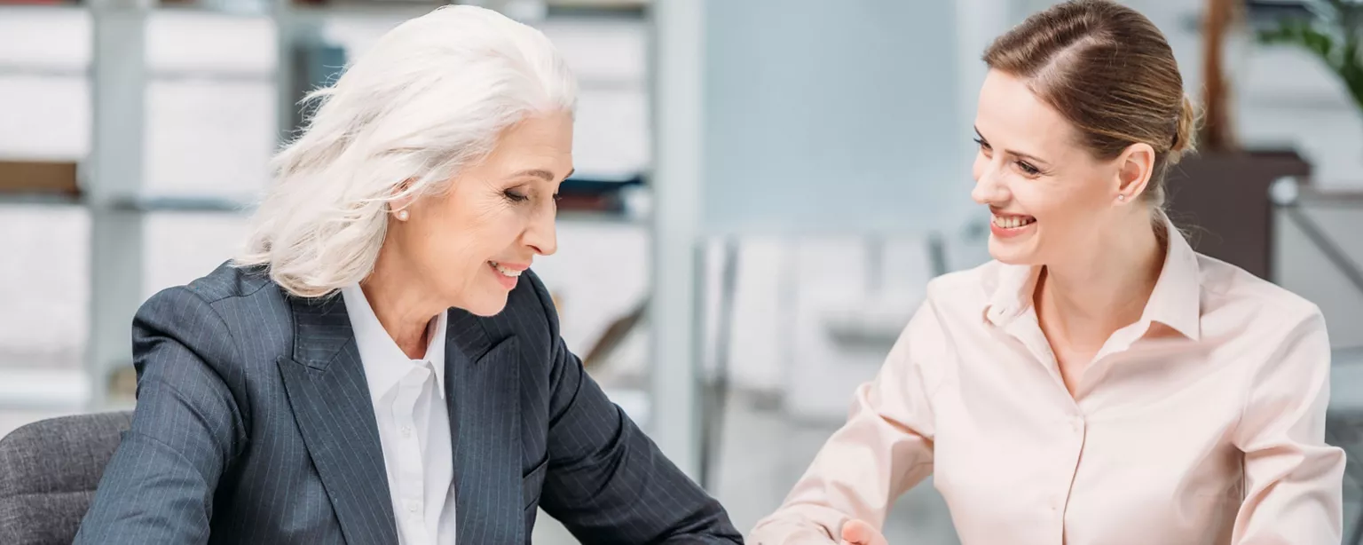 An older professional woman in a business suit engages in a friendly discussion with a younger female colleague, both smiling as they collaborate in an office.