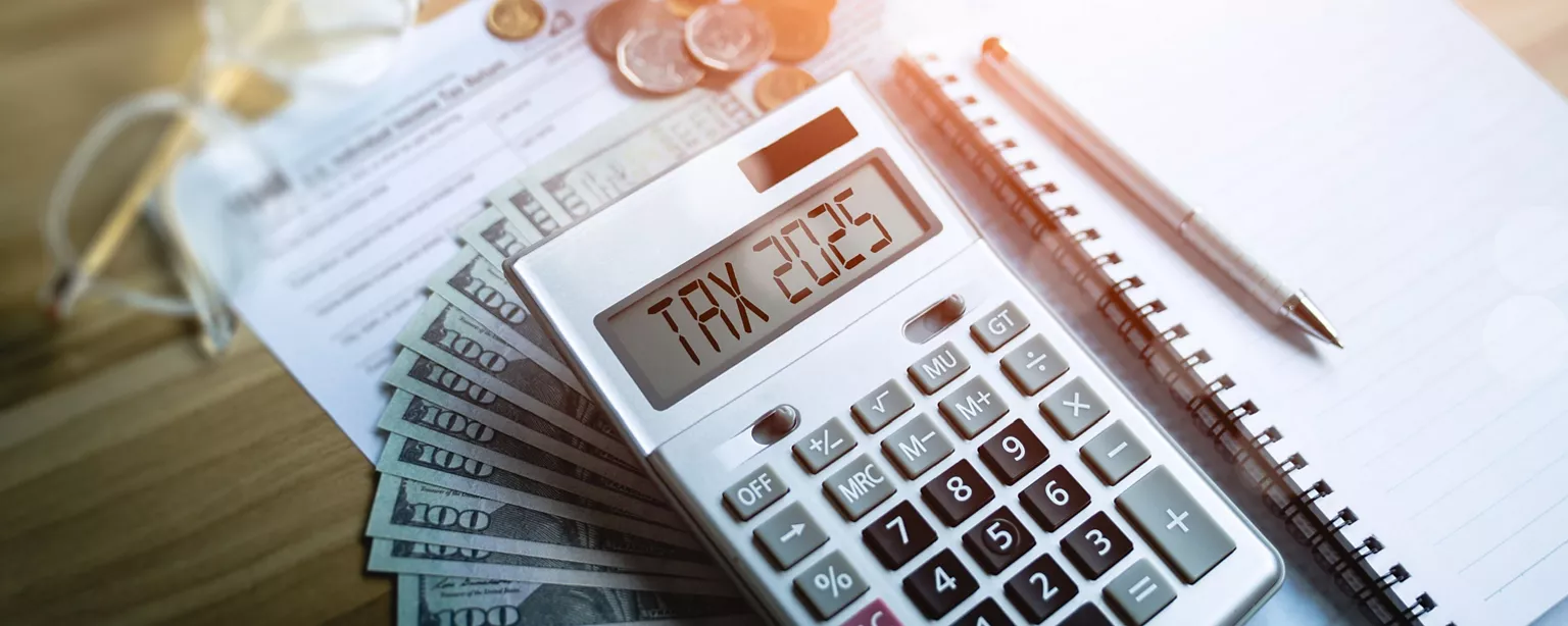 A calculator with the words Tax 2025 shown on it, rests on top of cash next to a notebook with a pen on it. 