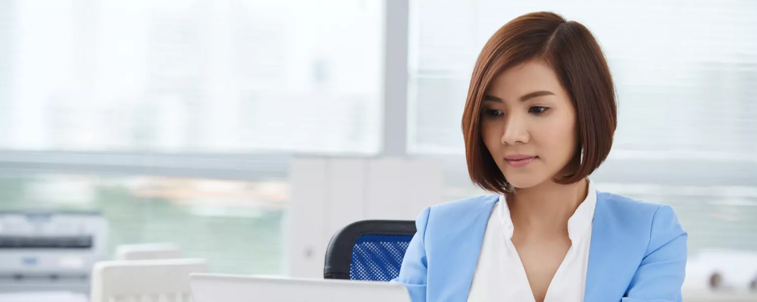 Young woman in a blue jacket working on a laptop in a corporate office.