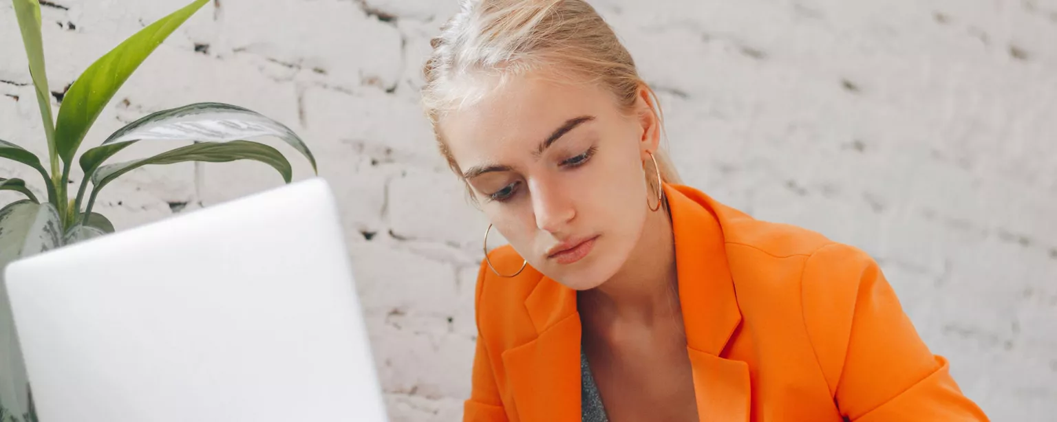 Blonde young woman in an orange blazer sitting in front of a laptop and writing in a notebook.