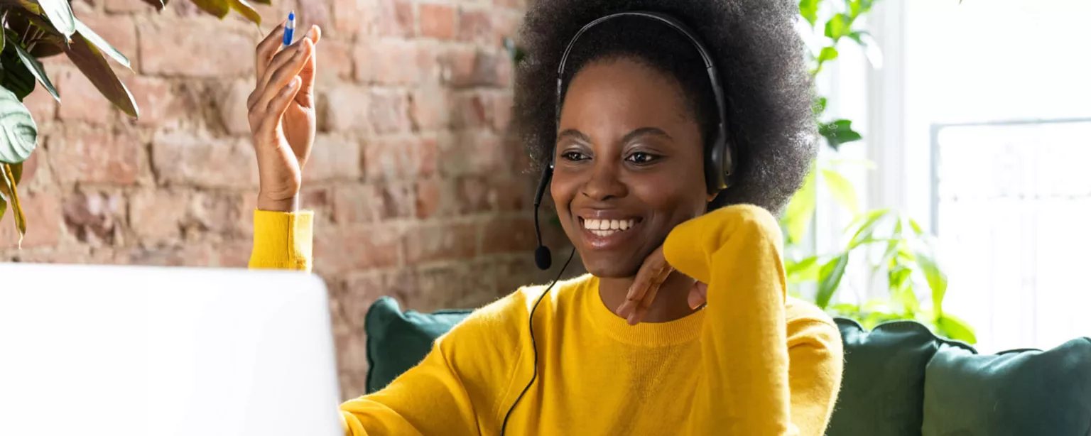 Young woman wearing headphones and a yellow sweater, sitting in front of a laptop and smiling while on a video call; the office is well-lit with plants, a brick wall and windows.