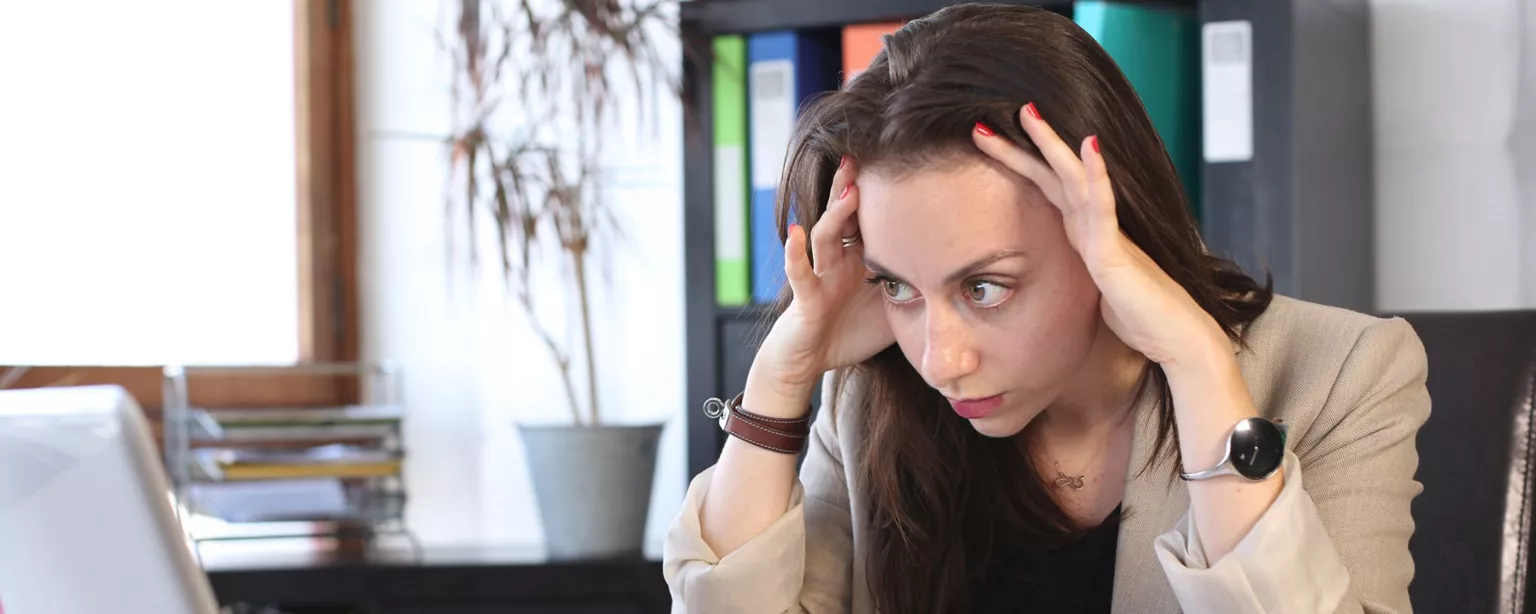 A finance and accounting professional sits at her desk, surrounded by financial charts and staring at a laptop screen. She is rubbing her temples and looking stressed.