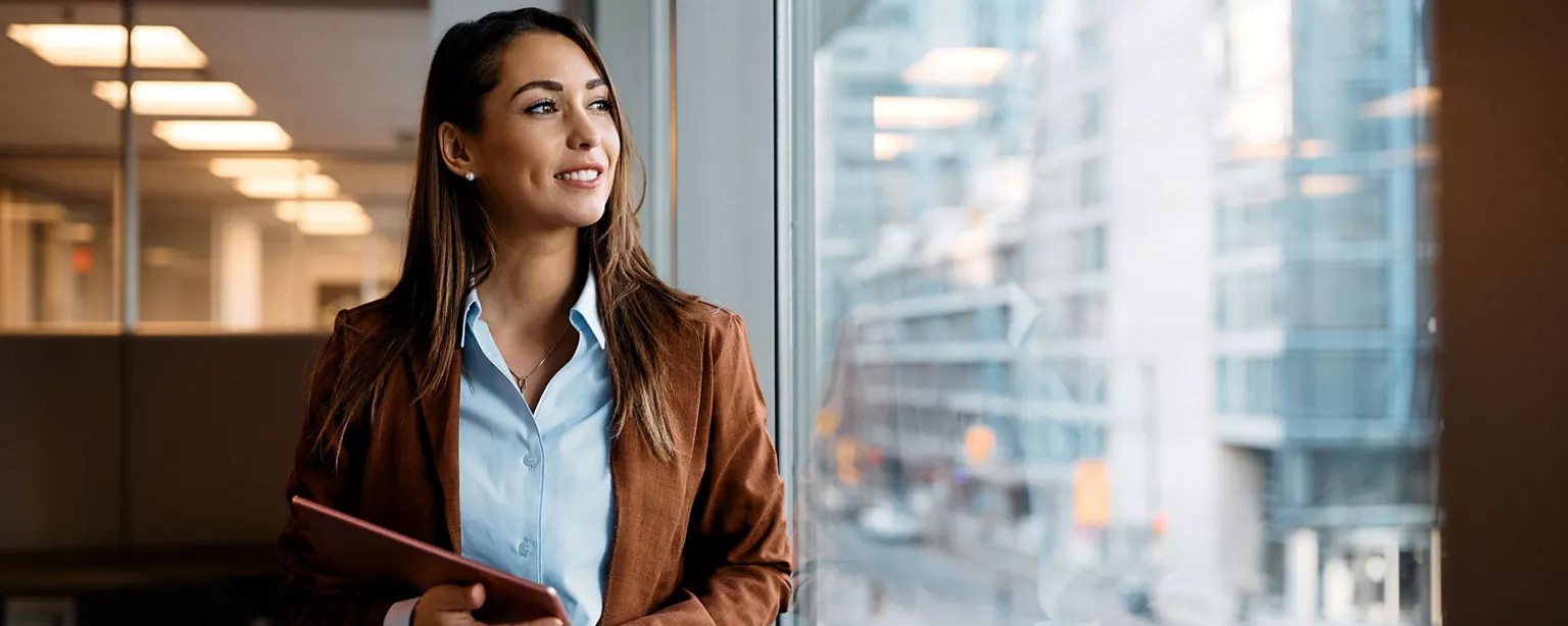 A smiling businesswoman holds a tablet while standing by a window in an office with a cityscape view in the background.