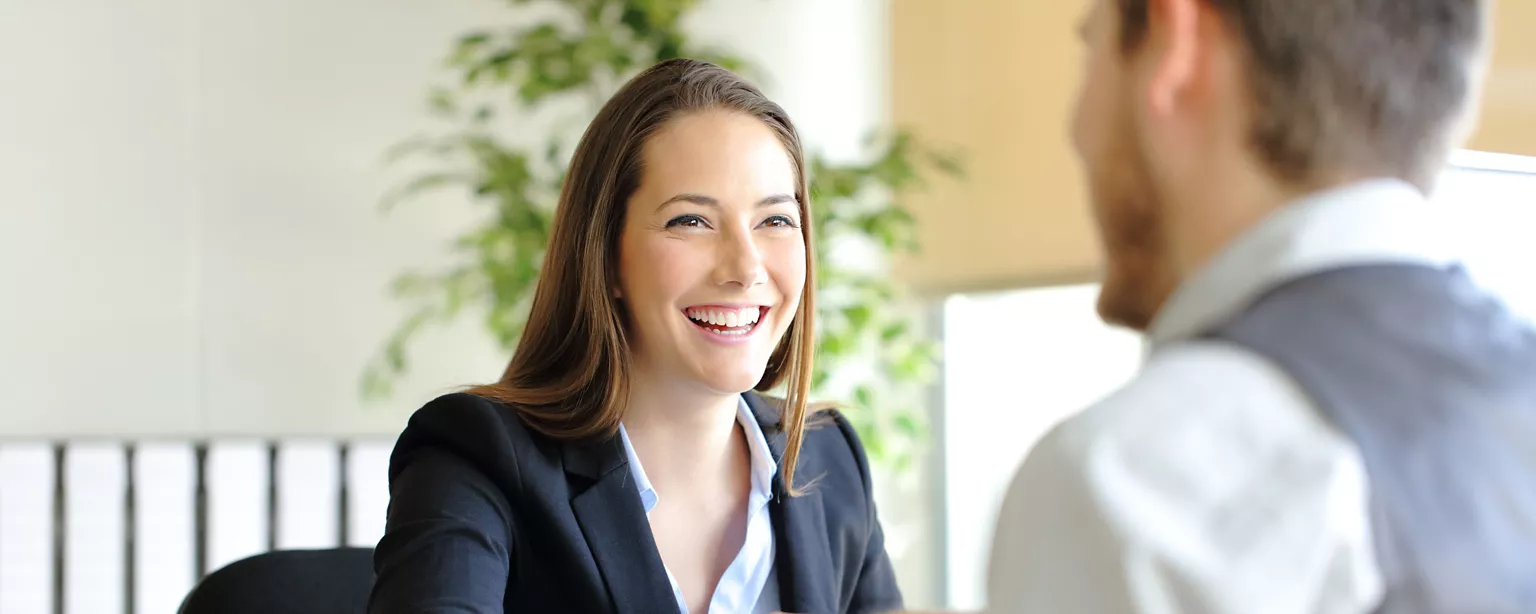 A woman in a business suit smiles warmly during a professional interview, seated across from a man at a desk.