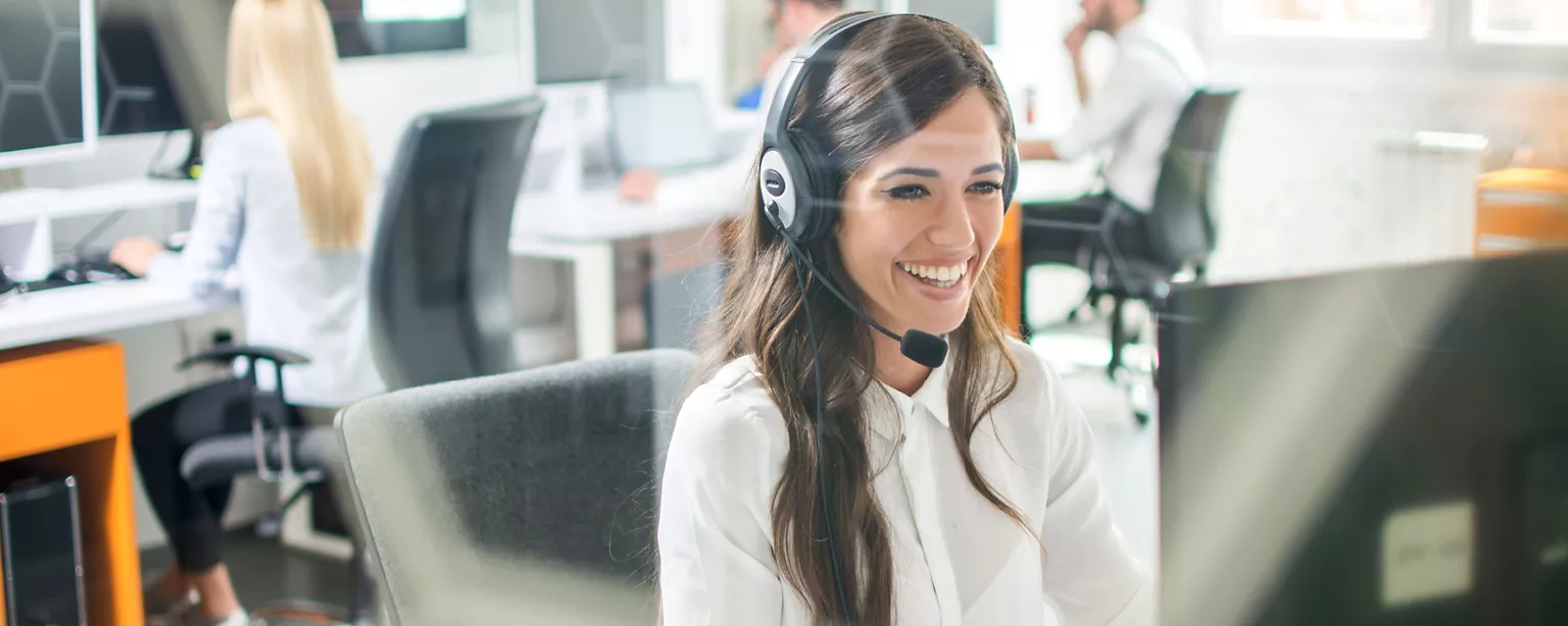 A woman wearing a headset smiles as she looks at her computer screen.