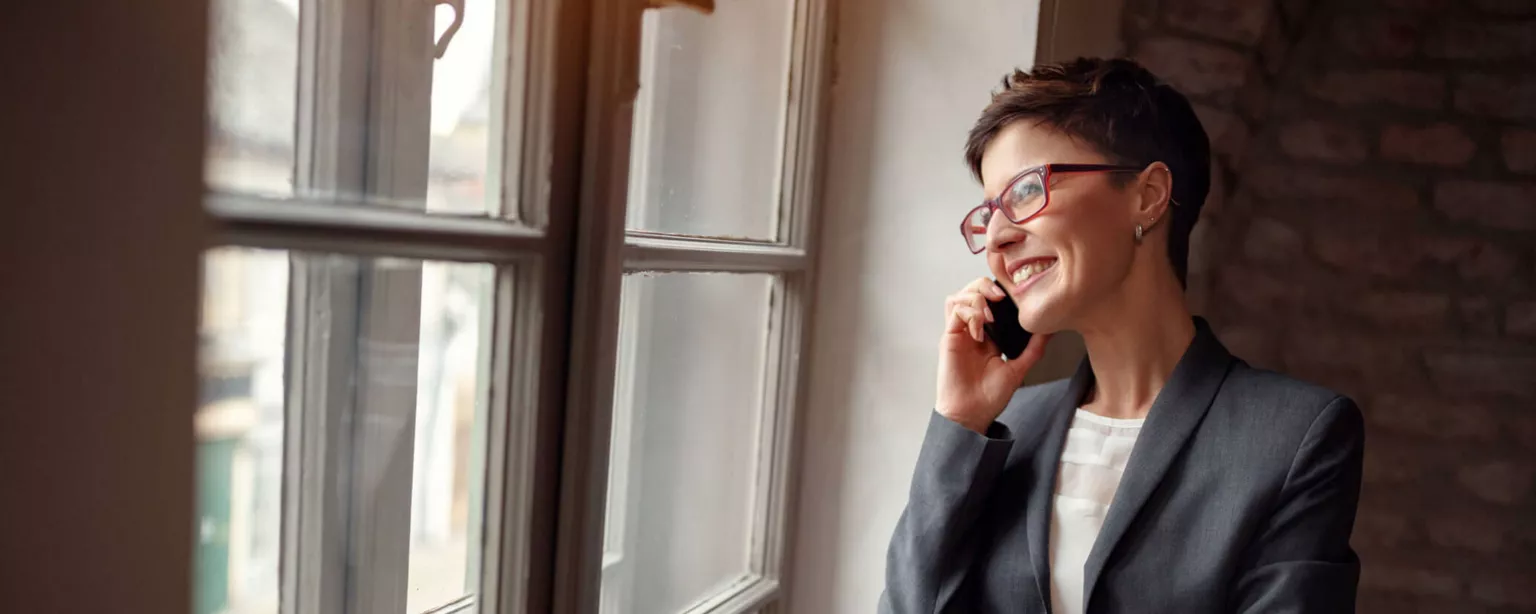 A woman executive talks on a mobile phone and smiles while looking out the window of her office.